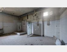 Verlassenes, kachelverkleidetes Zimmer mit beschädigten Wänden und Decken, defekten Rohren und einem zerbrochenen Fenster. Eine geöffnete Tür mit zersplittertem Glas und ein kleiner eingehobener Bereich sind sichtbar.