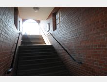 Treppe in einem überdachten Ziegelgang mit beidseitigen Metallhandläufen. Tageslicht fällt durch einen offenen Bogen am oberen Ende der Treppe, hinter dem zwei Personen zu erkennen sind.