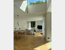 Ein modernes Wohnzimmer mit offenem Essbereich, Holzboden in Fischgrätmuster und großen Fensterfronten, die Tageslicht hereinlassen. Helle Möbel wie ein Esstisch mit Stühlen und eine Sofaecke dominieren den Raum, im Hintergrund ist ein großer Flachbildfernseher zu sehen, und durch die Fenster ist ein grüner Garten erkennbar.