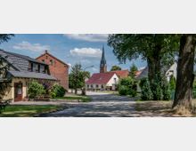 Straßenszene in einem Dorf mit gepflastertem Weg, umgeben von Einfamilienhäusern mit roten und grauen Dächern; im Hintergrund ragt der spitze Kirchturm einer Backsteinkirche in die Höhe, umgeben von Bäumen und Bewuchs.