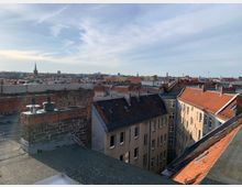 Blick von einem Dach auf eine städtische Wohngegend mit mehrstöckigen Gebäuden und roten Ziegeldächern. Im Hintergrund erstreckt sich die Skyline der Stadt mit vereinzelten Kirchtürmen und modernen Bauwerken unter einem blauen Himmel.
