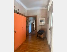 Flur in einer Wohnung mit Holzfußboden und cremefarbenen Wänden. Links steht ein orangefarbener Schrank, rechts ein brauner Sessel mit einer Tasche darauf, und im Hintergrund ist ein Durchgang zu einem weiteren Raum mit orientalischem Teppich sichtbar.