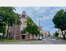 Straßenszene in einem Wohngebiet mit einem rosafarbenen Gebäude im Vordergrund, das eine dekorative Fassade und ein ovales Schild auf dem Dach trägt. Links und rechts der Straße stehen Bäume, während im Hintergrund weitere mehrstöckige Gebäude mit unterschiedlichen Fassadenstilen zu sehen sind.