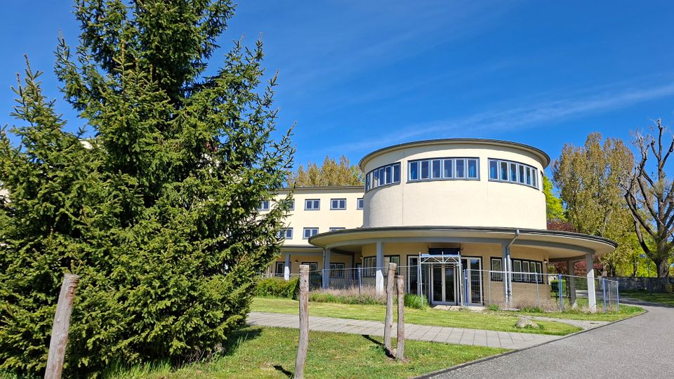 Modernes, hell verputztes Gebäude mit rundem Eckbau und umlaufendem Fensterband im Obergeschoss sowie überdachtem Eingangsbereich mit Säulen. Davor liegen Rasenflächen, ein gepflasterter Weg und ein Zaun, umgeben von Bäumen unter blauem Himmel.