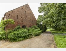 Backsteinbau mit einer Satteldachkonstruktion und kleinen kreuzförmigen Ziegelöffnungen, umgeben von dichtem Grün und Hecken. Ein sandiger Weg führt vorbei, flankiert von Bäumen und gepflegten Sträuchern.