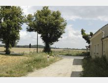 Ländliche Szene mit einem unbefestigten Weg, der an einem gelben Gebäude mit Regenrinne vorbeiführt. Links stehen Bäume und im Hintergrund sind Felder, Zäune und ein bewölkter Himmel erkennbar.