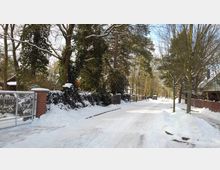 Eine verschneite Wohnstraße mit Bäumen, Zäunen und vereinzelt sichtbaren Häusern im Hintergrund. Der Gehweg und die Straße sind von Schnee bedeckt, während die Äste der Bäume leicht beschneit sind.
