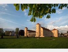 Barockschloss mit zwei runden Ecktürmen und rotem Dach, umgeben von einer gepflegten Grünanlage mit symmetrischen Beeten. Im Vordergrund hängen Kastanienblätter, der Himmel ist leicht bewölkt mit Abendstimmung.