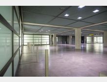 Offene Tiefgarage mit Betonboden, großen Säulen und einer Decke aus Metallgitter. Seitlich befinden sich Glaswände mit leichtem Milchglas-Effekt, und helle Beleuchtung erhellt den Raum gleichmäßig.