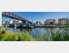 Fußgängerbrücke über einem Kanal in einer modernen Wohngegend mit mehrstöckigen, weißen Wohngebäuden im Hintergrund. Im Vordergrund befinden sich Gras und blühende Mohnblumen am Ufer, während mehrere Boote am Wasser liegen.