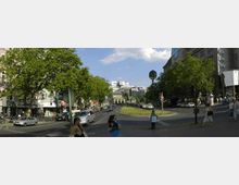 Straßenszene in einer Stadt mit breiten Gehwegen, Geschäften und Straßenverkehr. Im Hintergrund ist ein historisches Gebäude mit einem kleinen Grünbereich davor, umgeben von modernen Gebäuden und Bäumen.