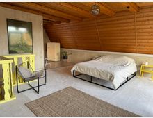 Schlafzimmer in einem Dachgeschoss mit schrägen Holzpaneelwänden und sichtbaren Holzbalken. Ein Bett mit weißer Bettwäsche steht auf einem hellen Teppich, daneben ein gelber Nachttisch; weitere Möbelstücke sind ein schwarzer Stuhl mit Kissen und ein gelbes Geländer. Im Hintergrund ist ein beige Kühlschrank, eine Pflanze und ein gerahmtes Bild an der Wand sichtbar.