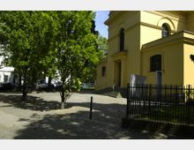 Gelbes Gebäude mit großen Rundbogenfenstern, vermutlich eine Kirche oder ein öffentliches Bauwerk, umgeben von Bäumen auf einem gepflasterten Platz. Im Vordergrund ist ein Metallzaun zu sehen, daneben eine kleine, mit Gras bedeckte Fläche.