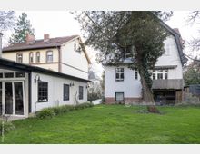 Gartenfläche mit Rasen und Bäumen, umgeben von zwei Wohnhäusern: einem modernen weißen Gebäude mit großen Fenstern links und einem älteren, weiß gestrichenen Haus mit roten Klinkerdetails rechts im Hintergrund. Die Umgebung ist ruhig und von Vegetation geprägt.