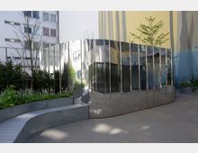 Moderner Außenbereich mit einer glänzenden, wellenförmigen Fassade aus Metall, die umliegende Gebäude und Pflanzen reflektiert. Im Vordergrund befinden sich bepflanzte Hochbeete und ein geschwungener Gehweg aus Holz und Beton.