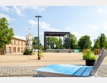 Eine offene Platzfläche mit einem modernen Metallpavillon als Bühne, umgeben von Bäumen. Links steht ein langes, historisches Ziegelgebäude, daneben ein dekorativer Turm aus Metallgittern, im Vordergrund eine blaue Sitzbank und ein großer Pflanzkübel.