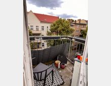 Ein kleiner Balkon mit Metallgeländer, einer schwarzen Sichtschutzmatte und verschiedenen Gegenständen wie Blumentöpfen, einer Wasserflasche und einer schwarzen Kunststoffstuhllehne. Im Hintergrund sind Wohngebäude mit roten Ziegeldächern und Bäume zu sehen, unter einem bewölkten Himmel.
