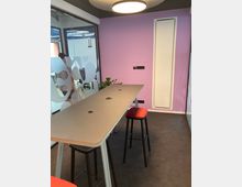 Moderner Besprechungsraum mit einem hohen Tisch und zwei roten Hockern, lila gestrichener Wand, gläserner Trennwand mit milchigen Mustern und dunklem Teppichboden. Eine runde Deckenleuchte spendet Licht, und an der Wand befinden sich Steckdosen.