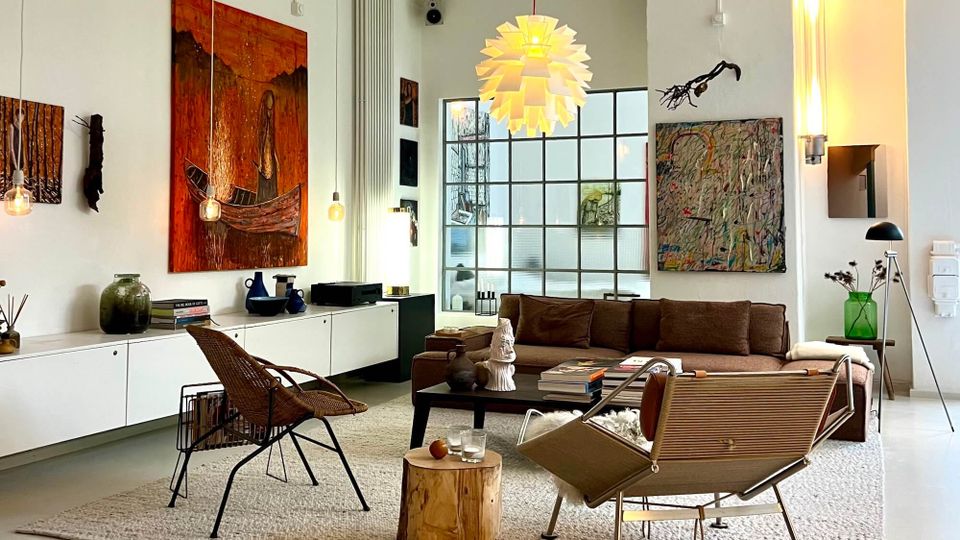 Modern living room with white walls, a large gridded window, and contemporary art pieces above a low white sideboard and brown sofa. A sculptural pendant light hangs over a rug with mixed lounge chairs and a coffee table, with minimalist floor lamps and decorative objects throughout.