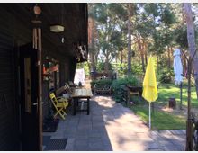 Außenbereich eines dunklen Holzhauses mit überdachter Terrasse, ausgestattet mit einem Holztisch, bunten Stühlen und Dekorationen. Im Hintergrund ist ein grüner Garten mit Bäumen, Sonnenschirmen und Sitzmöbeln zu sehen.