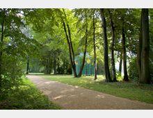 Ein Parkweg führt durch eine grüne, von Bäumen gesäumte Landschaft. Im Hintergrund steht ein kunstvoll gestalteter Pavillon mit grün-goldener Verzierung, umgeben von Rasen und Schatten spendenden Bäumen.