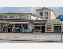 Straßenszene mit einem kleinen Einkaufszentrum, bestehend aus einem flachen Gebäude mit gelblichen Fassadenfliesen. Vor den Schaufenstern hängen gestreifte Markisen, darunter befinden sich Kleiderständer und Schaufensterauslagen, die Bekleidung präsentieren. Neben einem Geschäftseingang ist ein asiatisches Restaurant mit der Aufschrift „Orchidee Asia Küche“. Im Hintergrund steht ein mehrstöckiges Wohngebäude aus hellgelbem Putz. Rechts ist ein Schild für einen Behindertenparkplatz zu sehen.