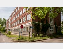 Vierstöckiges Gebäude mit roter und grauer Fassade, großen Fenstern und Fassadenteilen, die mit Efeu bewachsen sind. Im Vordergrund befinden sich ein Rasenstreifen, ein Zaun, ein Einbahnstraßenschild sowie ein Verbotsschild für die Einfahrt.