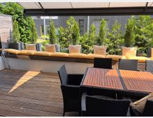 Sonnige Terrasse mit Holzboden, rattanähnlichen Sitzmöbeln und Holztischen. Im Hintergrund eine gepolsterte Sitzbank mit gelben Zierkissen vor einer Reihe von Grünpflanzen, die eine moderne Fassade aus Holz und Glas abschirmen.