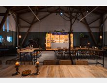 Großer Raum mit rustikalem Holzgebälk und einer zentralen Bar im Hintergrund, die von warmem Licht beleuchtet ist und von Pflanzen umgeben wird. Im Vordergrund stehen lange Holztische und Stühle, teilweise mit brennenden Kerzen dekoriert.
