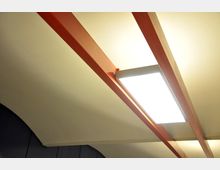 Decke mit roten Metallstreben und einer rechteckigen, hell leuchtenden Deckenlampe in modernem Stil. Die Umgebung scheint Teil eines Innenraums mit gebogenen Elementen zu sein.