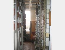 Schmaler Technikraum mit mehreren Reihen alter Telefonvermittlungsanlagen und Kabeln an den Wänden. Im Hintergrund befindet sich ein Fenster mit teilweise heruntergelassenem Vorhang, darunter ein Heizkörper und ein Stuhl.