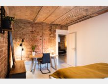 Schlafzimmer mit Sichtmauerwerk aus roten Ziegeln und einer gemauerten Gewölbedecke. Im Raum stehen ein Schreibtisch mit Stuhl, eine Tischlampe, eine Zimmerpflanze und links ein Sideboard; eine offene Tür führt zu einem weiteren Raum.