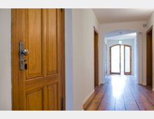 Holzverkleideter Flur mit einer hellen Holztür im Vordergrund, ausgestattet mit einem modernen Türschloss und Schlüssel. Der Flur besitzt helle Wände, einen Holzboden und führt zu einer doppelflügeligen Glastür am Ende, durch die Tageslicht einfällt.