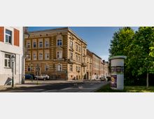 Straßenecke in einer Wohngegend mit einem gelblichen Altbau mit reich verzierten Fensterrahmen und Ziegelmustern. Im Vordergrund stehen eine Litfaßsäule mit Plakaten sowie eine grüne Laterne, daneben ein Gehweg mit Bäumen und parkenden Autos.