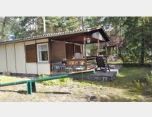 Kleines Ferienhaus aus Holz mit hellen Wänden und braunen Fensterläden, einer überdachten Terrasse mit Sitzgelegenheiten, umgeben von Bäumen und Wiesen in einer naturbelassenen Umgebung.