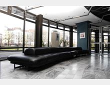 Innenansicht eines modernen Gebäudes mit bodentiefen Glasfenstern, die den Blick auf eine städtische Straße mit Bäumen und Gebäuden freigeben. Im Vordergrund befinden sich schwarze Ledersofas auf einem Boden aus grauem Marmor.