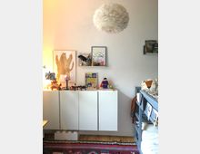 Ein Kinderzimmer mit einer weißen Wand und einer hängenden, federartigen Deckenlampe. An der Wand befindet sich ein weißes Sideboard mit Dekorationen, Spielzeug und Bilderrahmen, daneben steht ein dunkelgraues Etagenbett mit einer Wimpelkette. Ein bunter Teppich bedeckt den Holzboden.
