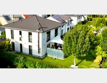 Modernes zweistöckiges Einfamilienhaus mit heller Fassade, einem dunkelgrauen Walmdach und großen Fenstern. Vor dem Haus befindet sich ein gepflegter Garten mit Rasen, Hecken und Bäumen, sowie eine Terrasse mit Sonnensegel und Glasabtrennung.