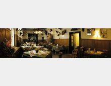 Gemütlicher Gastraum mit Holztäfelung an den Wänden, dekoriert mit ausgestopften Tieren und Jagdmotiven. Die Tische sind mit grünen Tischdecken, Blumen und Deckservietten geschmückt, und an der Decke hängen rustikale Lampen.