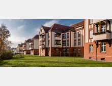 Mehrstöckige Wohngebäude im historischen Stil mit roten Backsteinfassaden und weißen Putzflächen, teilweise mit Balkonen. Im Vordergrund befindet sich eine Rasenfläche, die von Bäumen und niedrigen Büschen gesäumt ist.