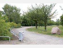 Ein Spazierweg in einem Park mit grasbewachsenen Flächen, Bäumen und Büschen, die den Weg säumen. Im Vordergrund steht ein großer Felsblock sowie eine mit Graffiti versehene Mülltonne, während im Hintergrund ein Jogger auf dem Weg zu sehen ist.