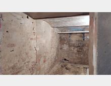 Ein kleiner, leerer Kellerraum mit freiliegenden, teils beschädigten Ziegelwänden und einer niedrigen Betondecke. Der Boden ist uneben, und an der rückwärtigen Wand sind Rohre sowie Spuren von Feuchtigkeit erkennbar.