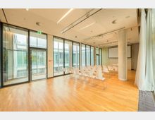 Heller Seminarraum mit großen bodentiefen Fenstern, Holzboden und weißem Mobiliar; in Reihen angeordnete Stühle, Leinwand und Projektor an der Decke. Fenster bieten Ausblick auf benachbarte moderne Gebäudefassaden.