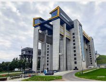 Hochmoderne Schiffshebeanlage aus Beton mit großen gelben Stahlträgern und Glasfenstern, umgeben von einer Grünfläche und einem asphaltierten Weg. Im Hintergrund ist eine ältere Stahlkonstruktion sichtbar, eingebettet in eine bewaldete Umgebung.