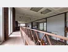 Großer Innenraum eines historischen, industriellen Gebäudes mit hohen Decken, einem Gittertragwerk und großen, rechteckigen Fenstern, die viel Tageslicht hereinlassen. Eine offene Galerie mit Holzgeländer verläuft entlang der Backsteinwände, und der Boden ist mit einem Schachbrettmuster aus Fliesen gestaltet.