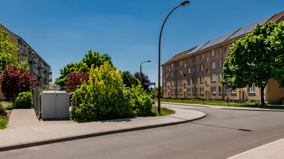 Kurvenreiche Wohnstraße in einem Siedlungsgebiet, gesäumt von mehrstöckigen Wohnblöcken mit Satteldächern. Breite Gehwege, Straßenlaternen sowie dichte Bäume und Sträucher prägen das Straßenbild.