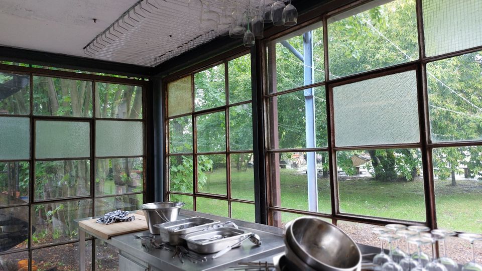 Ein Raum mit großen Fensterfronten aus Metallrahmen, die den Blick auf eine grüne Parklandschaft freigeben. Im Vordergrund stehen Küchengeräte, darunter Schüsseln, Tabletts und Gläser, auf Edelstahloberflächen.