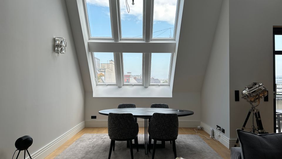 Moderner Essbereich in einem Dachgeschossraum mit schrägen Wänden und großem, mehrteiligem Dachfenster mit Blick auf die Stadt. In der Mitte stehen ein runder Tisch mit vier gepolsterten Stühlen auf einem Teppich, umgeben von hellem Holzboden und minimalistischer Beleuchtung.