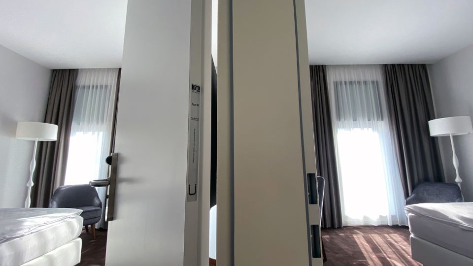 Ein schmaler Durchgang trennt zwei nahezu identische Hotelzimmer mit weißen Wänden, großen Fenstern mit halbtransparenten Vorhängen und dunklen Verdunkelungsgardinen. Die Zimmer enthalten jeweils ein Bett, einen Sessel und eine Stehlampe, mit Teppichboden in dunklen Farbtönen.