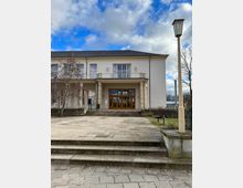 Hellbeiges zweistöckiges Gebäude mit Säulen am Eingang und großen Holz-Doppeltüren, die von breiten Fenstern flankiert werden. Vor dem Gebäude führen Stufen über einen gepflasterten Platz, umgeben von Grünflächen und einer Laterne auf der rechten Seite.